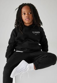 Felpa nera con scritte bianche, pantaloni sportivi neri coordinati e scarpe bianche. Il bambino ha i capelli ricci lungo le spalle. Look semplice e casual.