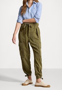 Olivgrön cargo-byxor med hög midja och dragsko, som har flera fickor och elastiska muddar, tillsammans med en ljusblå skjorta med knappar.