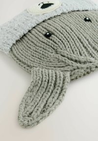 Juguete de peluche gris tejido en forma de ballena, con un gorro difuso de color gris claro, ojos de plástico negro y un detalle facial blanco bordado.