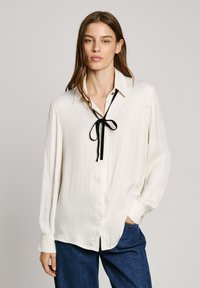Blusa crema con colletto e cravatta nera. Maniche lunghe con polsini bottonati. Tessuto liscio con una vestibilità ampia. Abbinata a jeans blu.