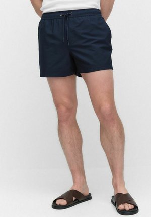 Shorts de bain bleu marine en tissu texturé, avec une taille élastique et un cordon de serrage ajustable. Portés avec des sandales à brides croisées marron.