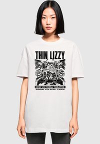 Bela oversize majica s črnim grafičnim tiskom "Thin Lizzy" in gledališkim logotipom ter podrobnostmi na dnu. Kratke rokave, okroglo izrezana.