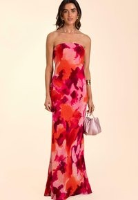 Robe maxi sans bretelles avec un imprimé aquarelle vibrant rouge et rose, tissu lisse, corsage ajusté, jupe fluide, associée à un petit sac à main rose.