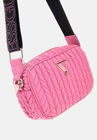 Rosa kviltad crossbody-väska med texturerat läder, guld hardware, logodetalj och justerbar svart rem med ordet "GUESS."