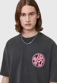 Ciemnoszara, oversize koszulka z krótkim rękawem, z okrągłym, różowym logo graficznym na lewej piersi. Gładka bawełniana faktura.