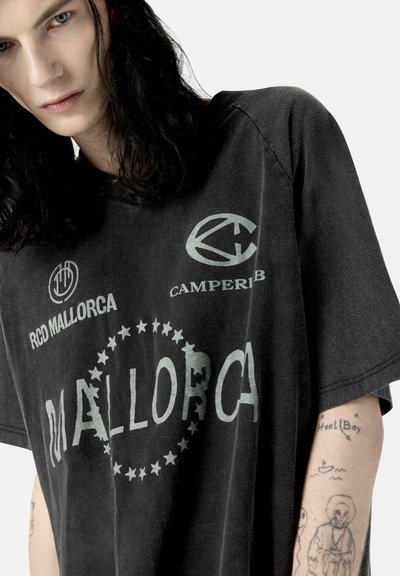 Personne aux longs cheveux foncés portant un t-shirt noir délavé avec "Mallorca" et des logos imprimés à l'avant et des tatouages visibles sur le bras.
