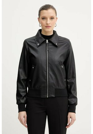 Donna che indossa una giacca nera in ecopelle con zip, colletto con bottoni a pressione e tasche laterali, abbinata a pantaloni neri, in piedi davanti a uno sfondo bianco.