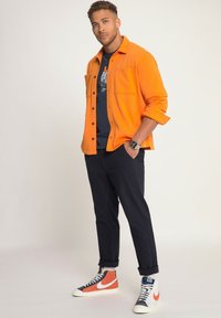 Camisa de botones naranja sobre una camiseta navy, combinada con pantalones oscuros y zapatillas altas multicolores. Presenta puños enrollados y bolsillos.