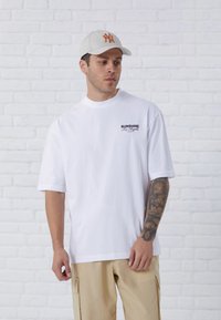 White short-sleeve t-shirt with a printed logo; paired with beige pants and a light gray cap featuring an orange embroidered design.