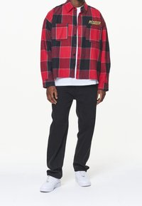 Czerwona i czarna flanelowa koszula w kratę z dwoma kieszeniami na piersi i haftem "PECADOR" w żółtym kolorze. Noszona na białej koszuli, zestawiona z czarnymi spodniami.