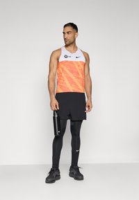 Sporditop oranž ja valge geomeetrilise mustriga, koos mustade lühikeste pükste ja legginsitega. Käes on must veepudel.