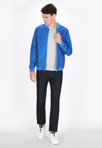 Giacca bomber blu con motivo a foglie testurizzato, t-shirt grigia chiara, jeans scuri e sneakers bianche. Chiusura frontale con zip e polsini a costine.