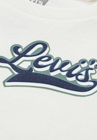Vit bomullströja med en framträdande blå broderad "Levi's"-logotyp med grön kant och mörkblå detaljer, texturerad design.