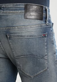 Jeans de denim azul con un acabado desgastado, con un parche de cuero en la cintura y bolsillos traseros estándar con detalles de costura.