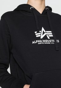 Sudadera negra con capucha y cordón, con un logo blanco y rayas en el pecho. Fabricada con tela suave y con un bolsillo tipo canguro.