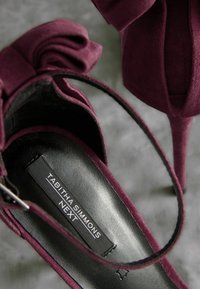 Sandale à talons hauts en suède bordeaux avec un détail nœud, intérieur en cuir noir et une étiquette de marque visible à l'intérieur. Design élégant et épuré.