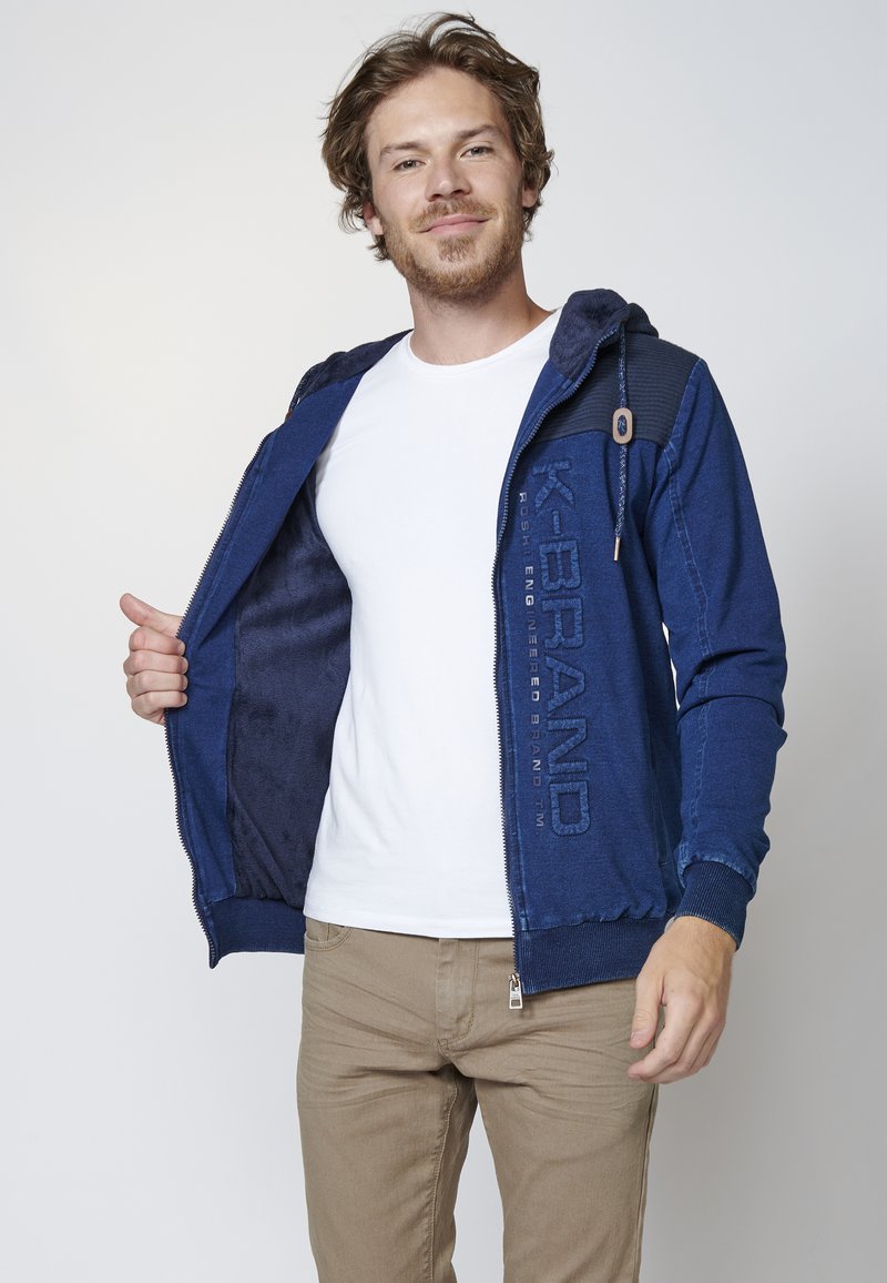 Blue zip-up hoodie with a soft interior, featuring a textured patch on the shoulder and embossed logo on the front. White t-shirt underneath.