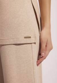 Beżowy sweter o lekko luźnym kroju, z bocznym rozcięciem i małą metalową metką z logo na dolnym brzegu. Gładka tekstura i prosty design.