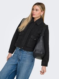 Femme blonde portant une veste en laine noire et un jean bleu, portant un sac à bandoulière en cuir noir, regardant sur le côté devant un fond uni.