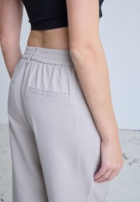 Personne portant un pantalon beige clair à taille élastique avec une poche arrière unique, associée à un haut court noir, debout de profil.