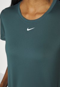 Camiseta atlética en verde azulado oscuro con una textura suave, cuello redondo y mangas cortas. Presenta un pequeño logo blanco de Nike en el pecho.