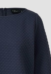 Marineblaues, strukturiertes Oberteil mit gestepptem Rautenmuster, Rundhalsausschnitt und Reißverschluss am Rücken. Das Material wirkt weich und formstabil.