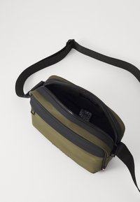 Olivengrüne Tasche mit einem schwarzen Reißverschluss und Tragegurt. Strukturierte Oberfläche, rechteckige Form, die einen teilweise geöffneten Reißverschluss und Details des Innenstoffs zeigt.