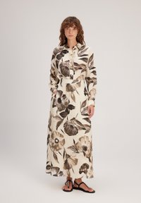 Robe longue à fleurs avec un fond beige et des motifs de fleurs brunes, un devant à boutons et des manches longues. Portée avec des sandales noires.