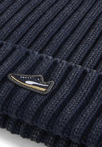 Gorra de punto acanalado en color marino, hecha de una mezcla de lana, con un patrón texturizado y un pequeño parche bordado en azul, blanco y dorado.