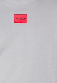 Sudadera gris claro con una textura acanalada y un parche rectangular rojo etiquetado con "HUGO" en el pecho.