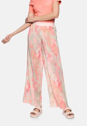 Frau trägt plissierte, weite Hosen mit rosa und beigem abstraktem Muster, korallenfarbenes Kurzarm-Top und sandfarbene Sandalen.