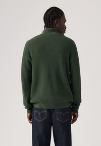 Žalias tarkuotas megztinis su aukštu apykakliu, derinamas su tamsiai mėlynais džinsais. Audinys atrodo minkštas ir jaukus, su laisva silueto forma.