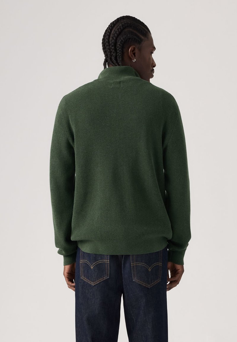 Grüner, gerippter Strickpullover mit hohem Kragen, kombiniert mit dunkelblauen Jeans. Die Textur wirkt weich und gemütlich, mit einer entspannten Silhouette.