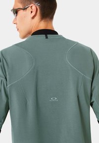 Mintgrünes Langarmshirt mit Rundhalsausschnitt und schwarzen Akzenten; verfügt über Nähdetails und das Oakley-Logo am unteren Rücken.