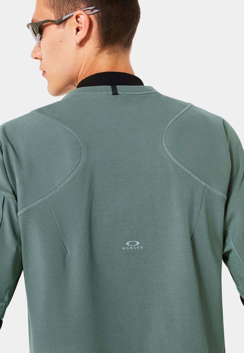 Chemise à manches longues vert menthe avec un col rond et des accents noirs ; présente des détails de coutures et le logo Oakley dans le bas du dos.