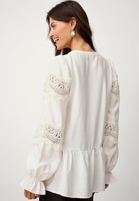 Weiße Bluse mit langen Puffärmeln, ausgestattet mit Spitzenakzenten an den Ärmel, einem gerafften Saum und einer strukturierten Oberfläche.