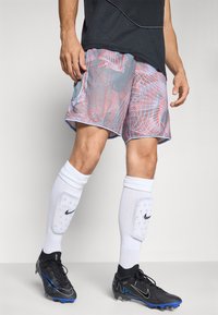 Mannlig utøver iført sports shorts med abstrakt mønster, hvite leggbeskyttere, hvite sokker og svarte Nike fotballsko, stående på et grått gulv.