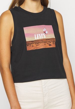 Kobieta ubrana w czarny bezrękawnik typu crop top z krajobrazem pustynnym, motylem i tekstem "LEVI'S" z przodu, w połączeniu z jasnymi spodniami.