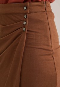 Fustă maro cu un material texturat, pliuri laterale și cinci nasturi decorativi negri pe talie. Închidere cu fermoar la spate.