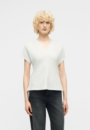 Persoon met kort krullend blond haar, een witte korte mouw V-hals top en donkere spijkerbroek, staand tegen een effen achtergrond.