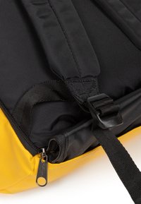 Detalj av en svart och gul ryggsäck som visar det strukturerade tyget, dragkedjans beslag, justerbara remmar och en snabbkopplingsspänne.