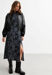 Schwarze Leder-Cropped-Jacke über einem dunkelgrauen Kleid mit schwarzen abstrakten Mustern, das einen Seitenschlitz hat, sowie kniehohen schwarzen Stiefeln mit Riemchen.