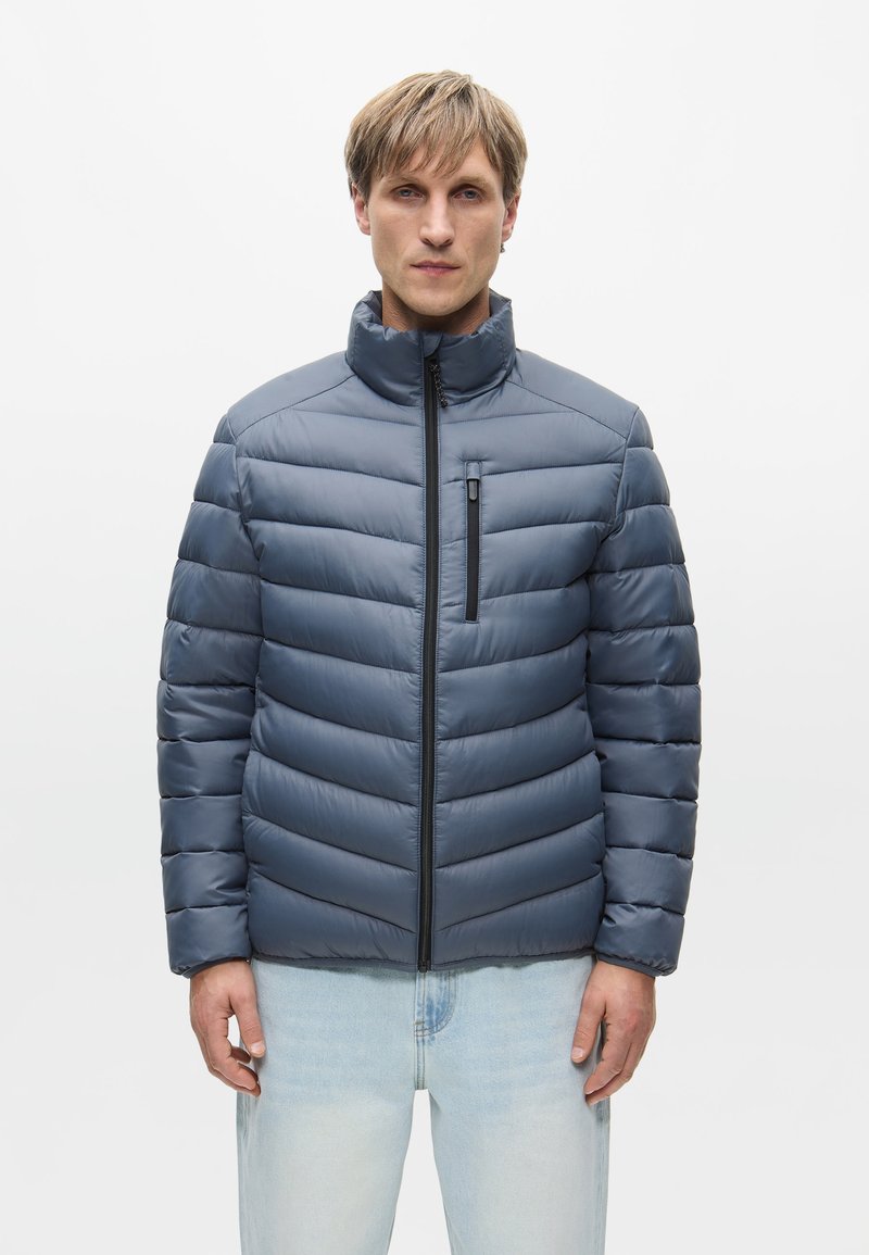 Férfi világoskék puffertalpú kabátban, magas gallérral és fekete cipzárral, világos színű farmerrel, sima háttér előtt állva.