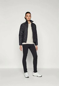 Melnas quiltētas jakas ar stāvošu apkakli, uzvilktas virs gaiši beidzona topa, apvienotas ar melnām legingām un baltām sporta kedām.