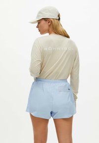 Shorts de color azul claro con cinturilla elástica y bolsillo trasero, combinados con una camiseta de manga larga beige con texto blanco. Gorro blanco llevado hacia atrás.