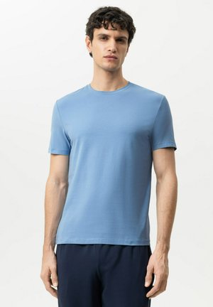 Man met donker krullend haar die een eenvoudige lichtblauwe T-shirt met korte mouwen en een donkere broek draagt, staand tegen een witte achtergrond.