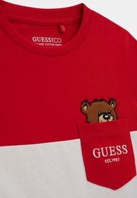 Punainen ja valkoinen orgaaninen puuvillapaita, jossa on karhupistoilu taskussa ja "GUESS"-logo punaisella ompeleella valkoisella paneelilla.