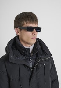 Giacca a vento nera con colletto alto e chiusura a zip, indossata sopra un dolcevita chiaro. Occhiali da sole neri quadrati con lenti oscurate.