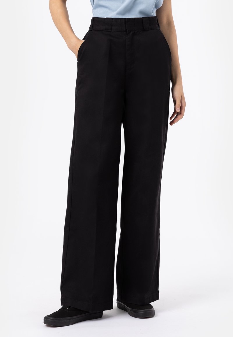 Svarta vida byxor i ett slätt, robust tyg, med hög midja och sidofickor, bärs med svarta skor.