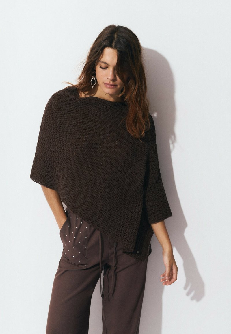 Poncho en tricot marron avec une coupe ample, un décolleté large et des manches mi-longues. Associé à un pantalon marron avec des pois blancs.
